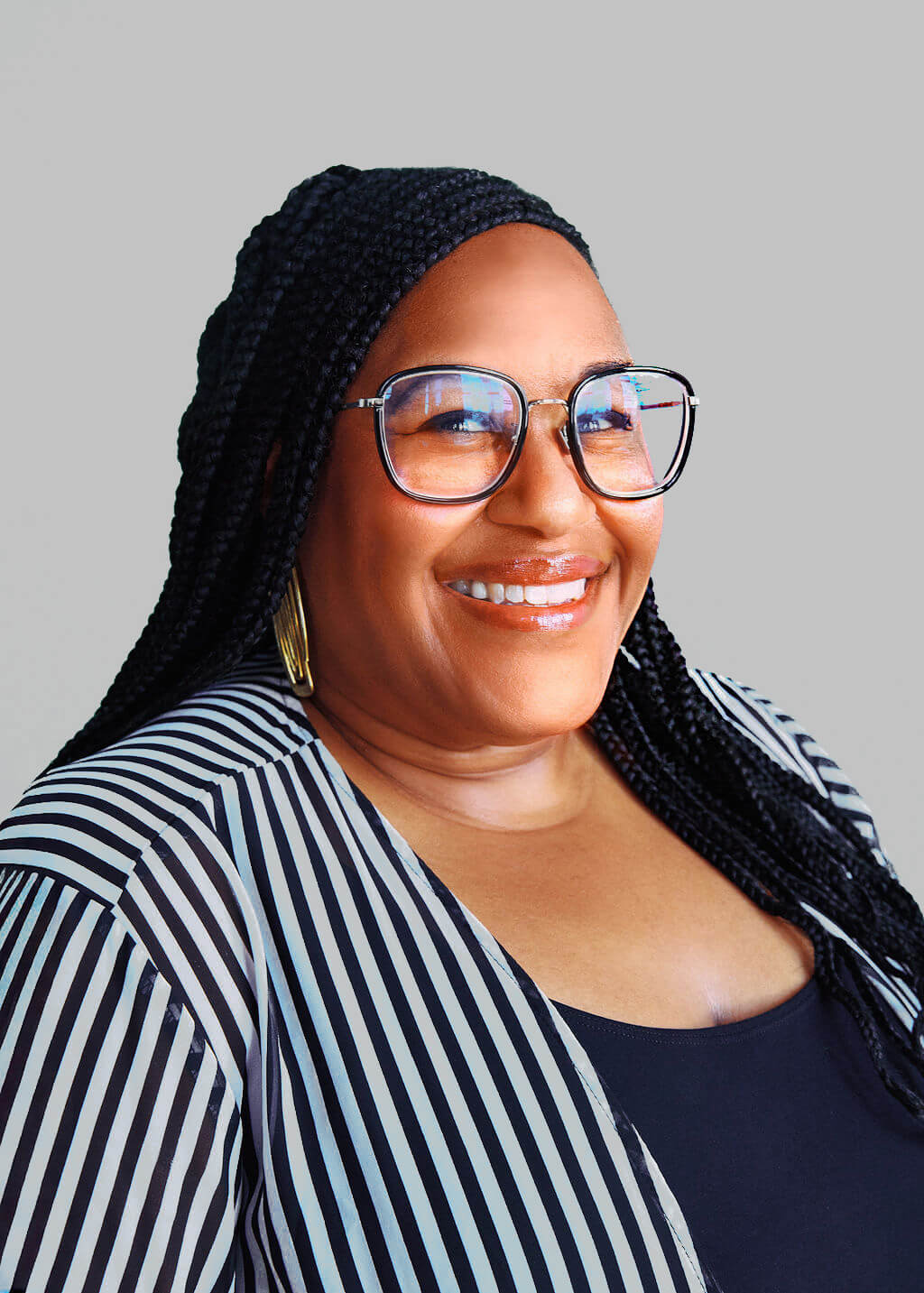 A woman with long braided hair, wearing glasses, gold earrings, a black top, and a black-and-white striped jacket, smiles against a plain light gray background—an effortlessly stylish look inspired by Abbré McClain.