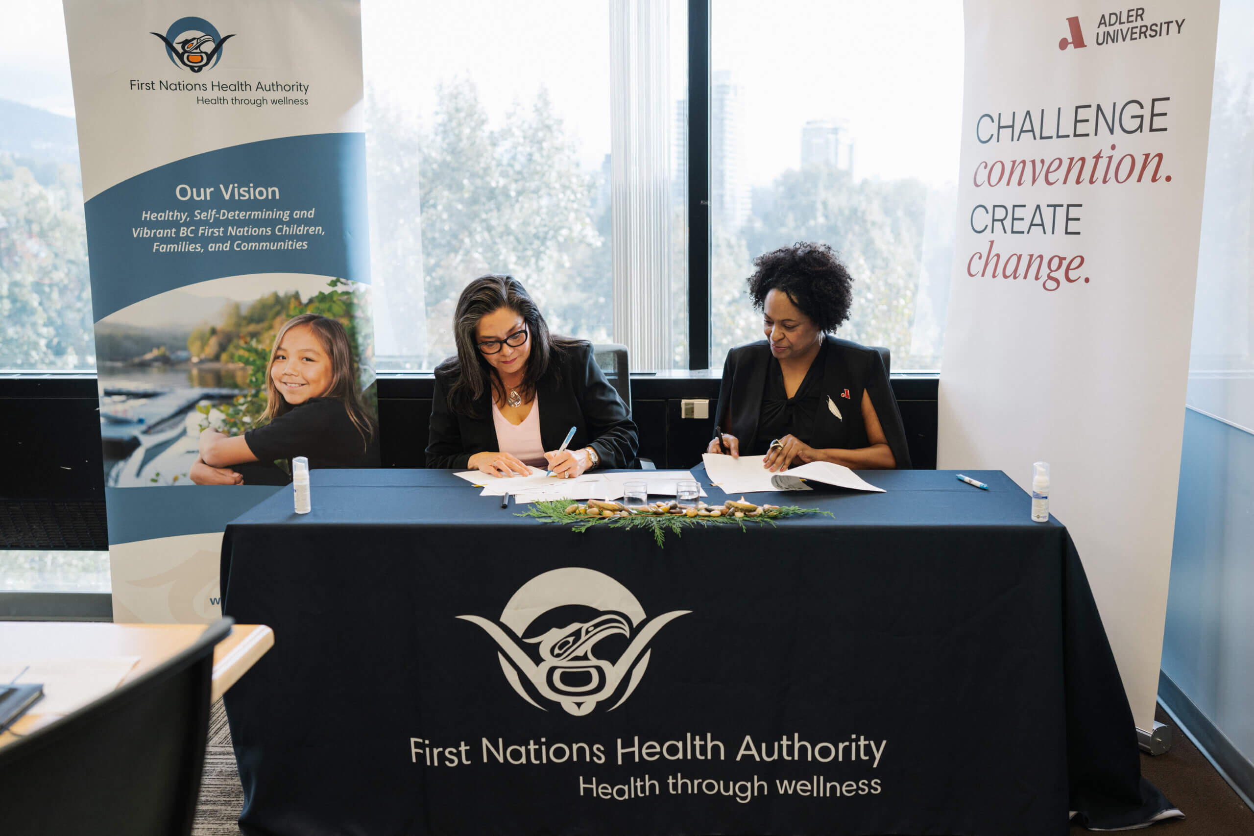 Monica McAlduff and Lisa Coleman, Ph.D., sitting at a table draped in a blue tablecloth with the FNHA logo.