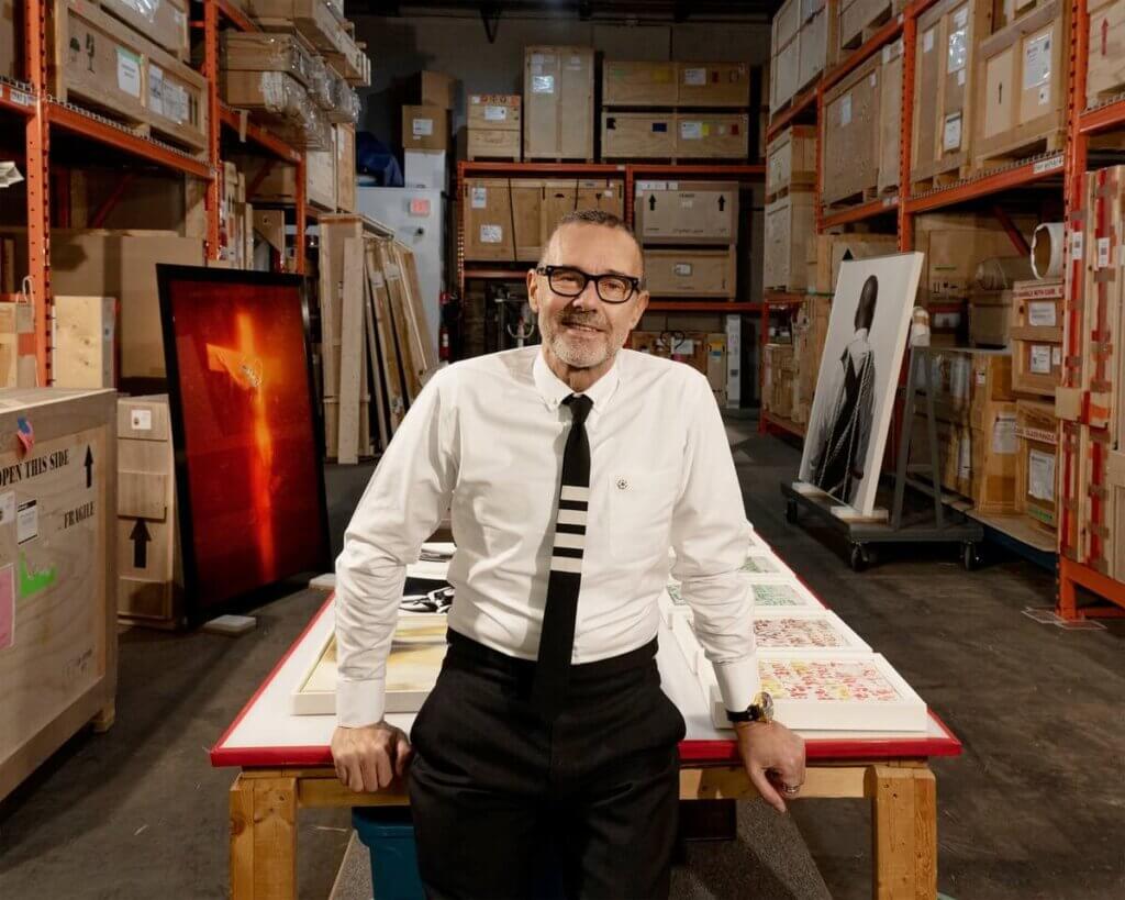 A man wearing glasses and a white shirt with a striped tie stands in a warehouse, leaning on a table with artwork and crates in the background—reminiscent of Adler University’s 2025 Commencement and its inspiring keynote speakers.