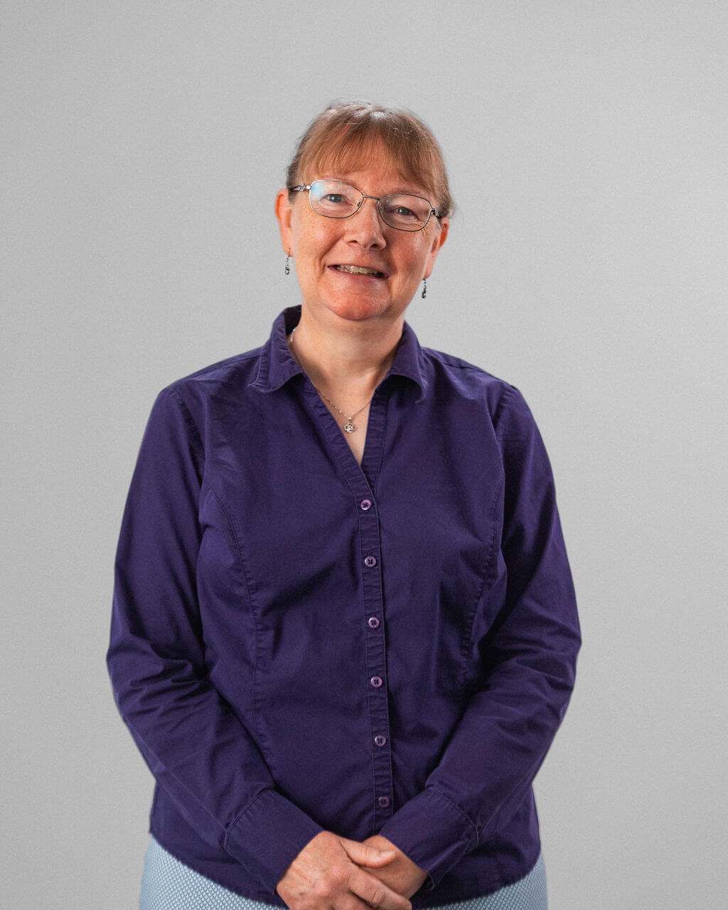 Debbie Clelland, a woman with short light brown hair and glasses, wears a purple button-up shirt as she stands in front of a plain light gray background, smiling with her hands clasped.
