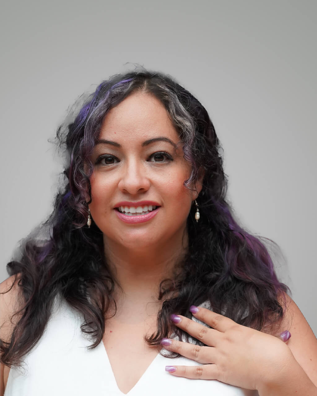 Flor Esquivel, a woman with long, wavy hair featuring purple streaks, smiles at the camera. She wears a white top, drop earrings, and sports matching purple nail polish.