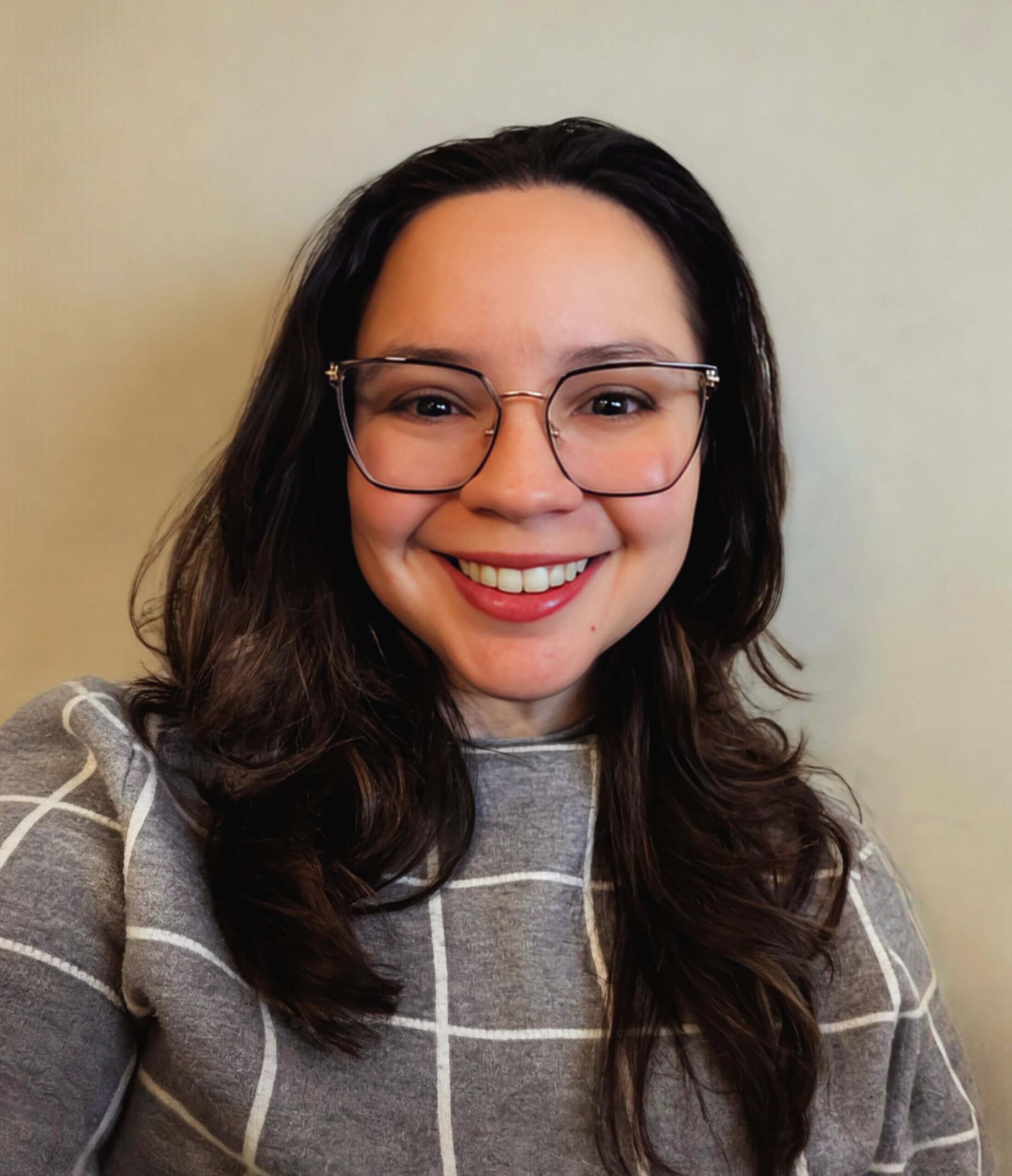 A woman with long brown hair, glasses, and a gray windowpane sweater smiles at the camera against a neutral background.