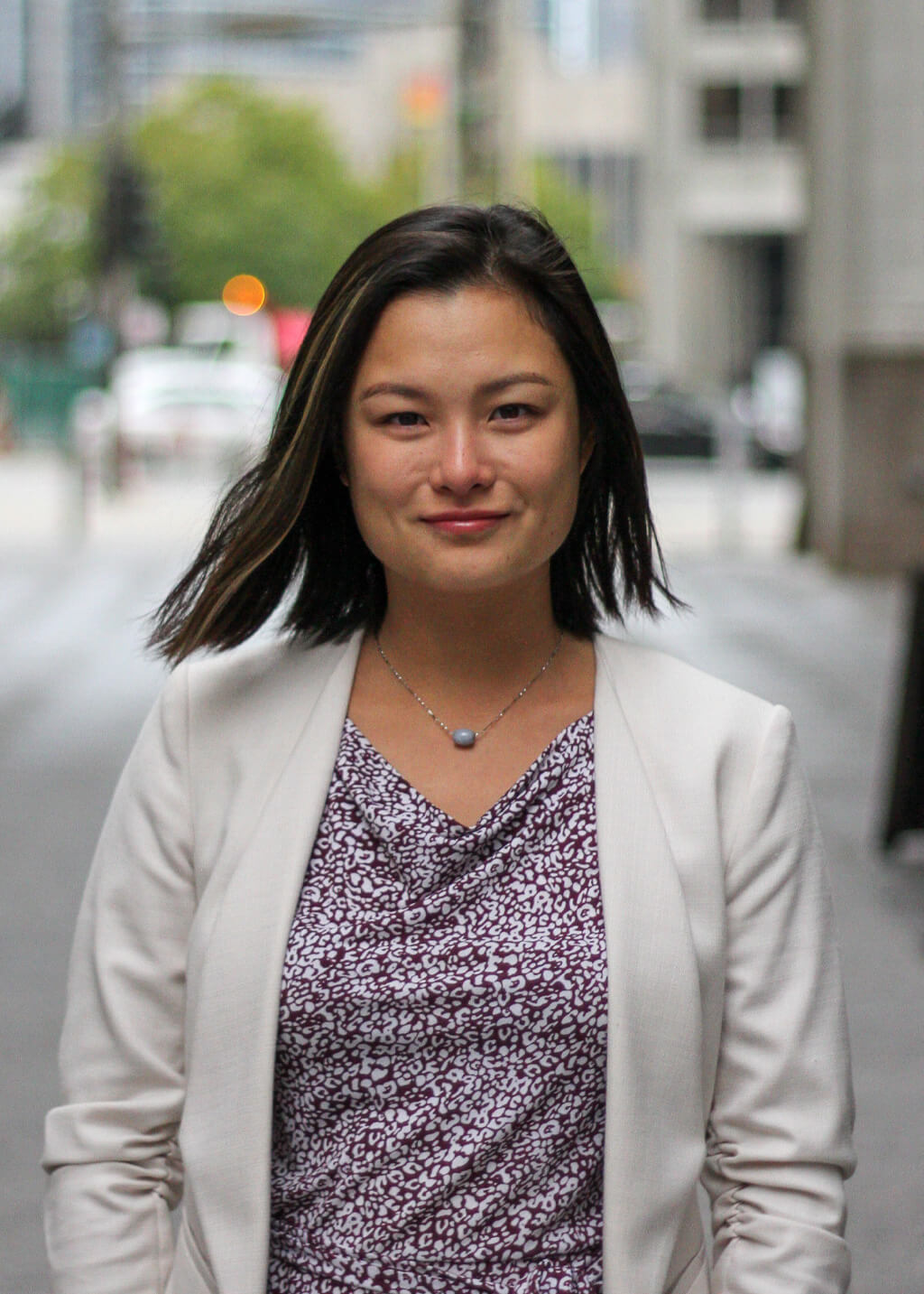 A woman with straight dark hair wearing a white blazer and patterned top stands outdoors on a city street, looking at the camera.