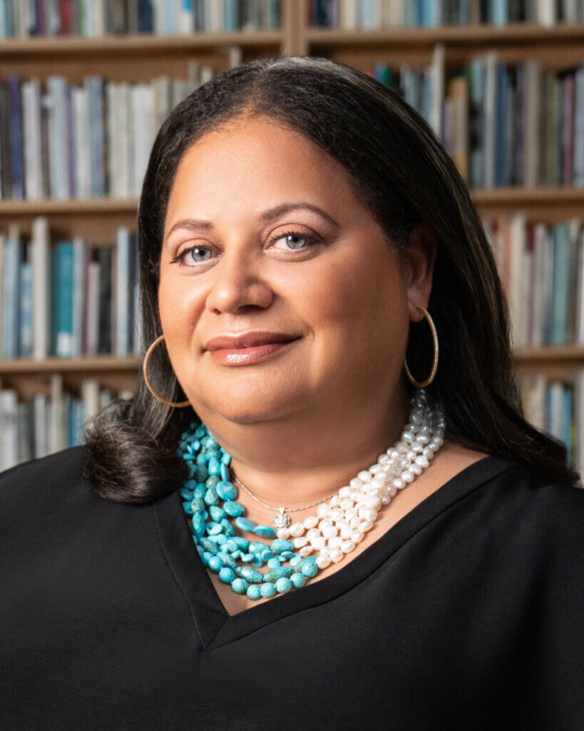 A woman with dark hair, wearing hoop earrings and layered turquoise and pearl necklaces, poses in front of a blurred bookshelf background—an inspiring choice among 2025 Commencement keynote speakers at Adler University.
