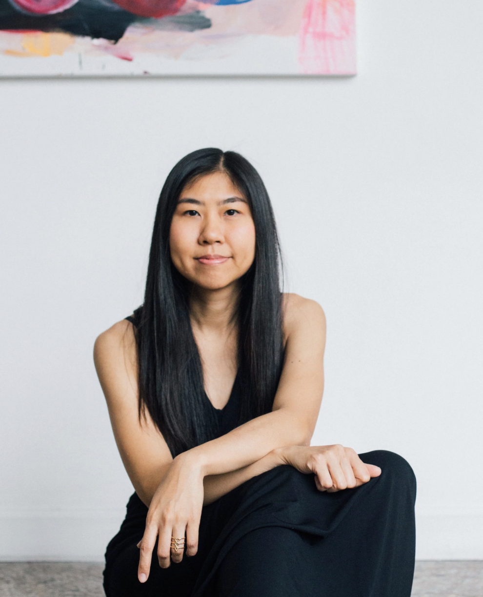 A woman with long black hair sits on the floor in a black dress, smiling slightly, with a colorful abstract painting on the wall behind her.