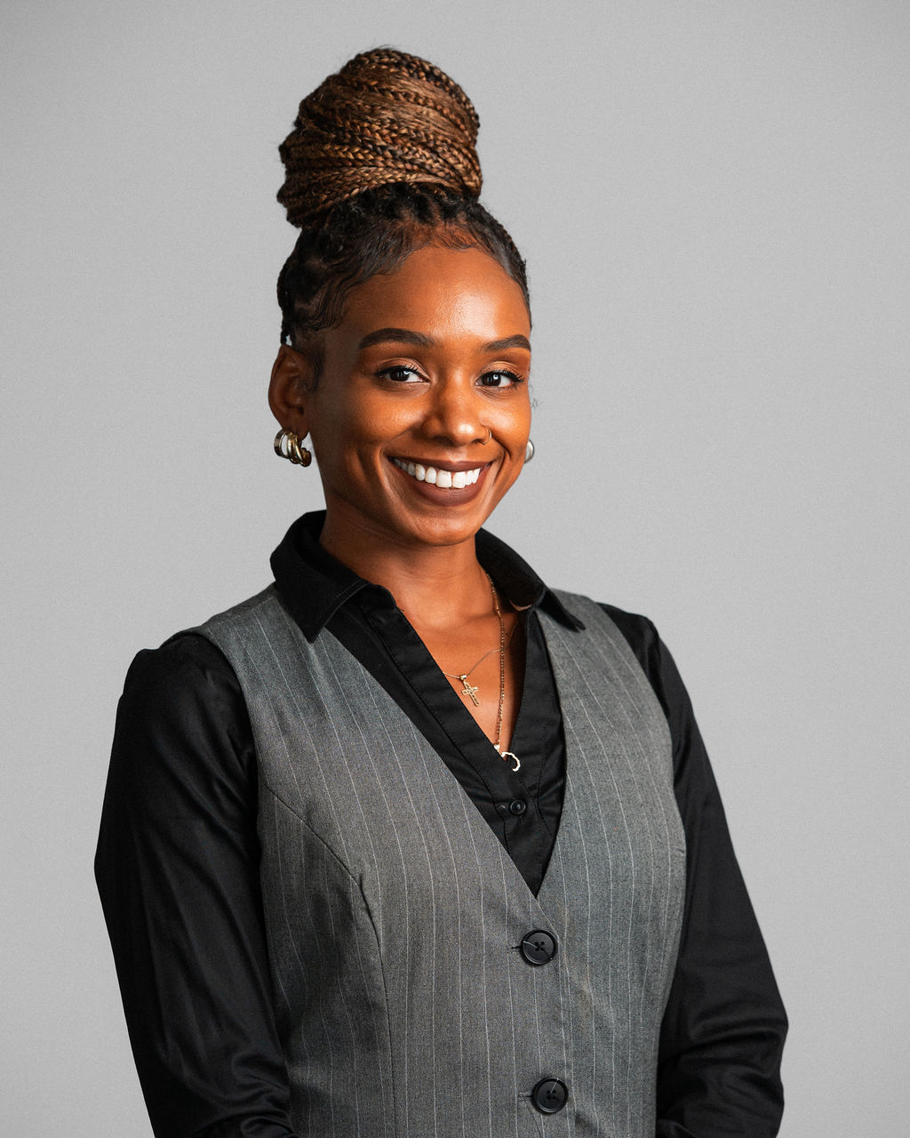 Dominique Barnes-Walker, with braided hair styled in a high bun, smiles at the camera in a black shirt and gray pinstripe vest against a plain gray background.