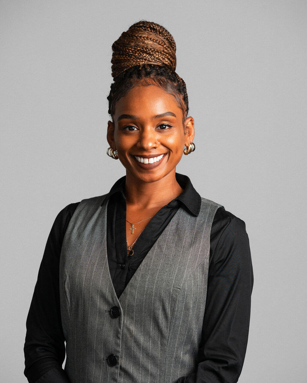 Dominique Barnes-Walker, a woman with braided hair in a high bun, wearing a black shirt and gray vest, smiles at the camera against a plain gray background.