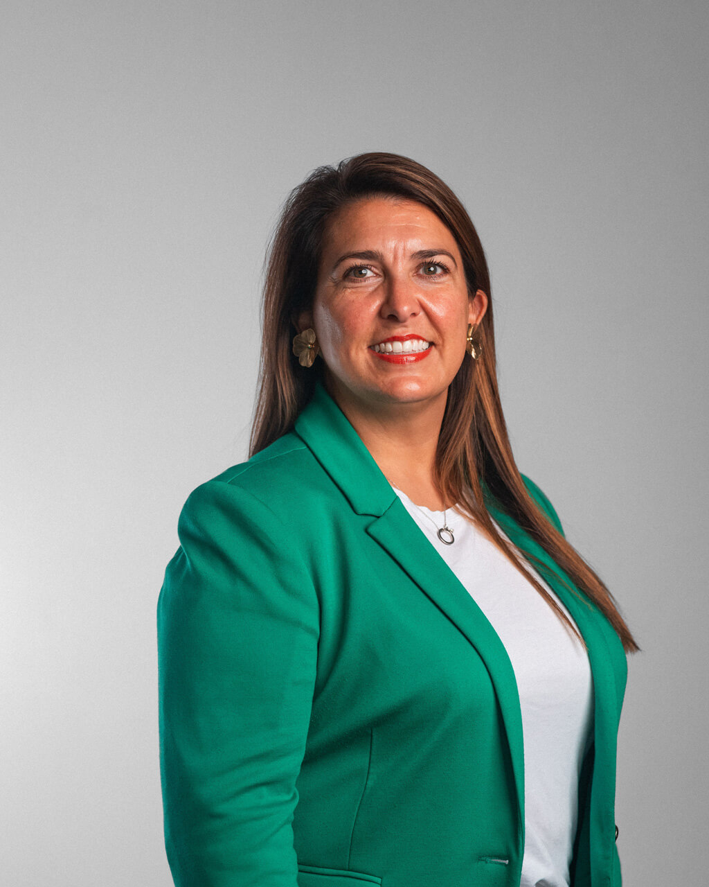Heather Miller, a woman with long brown hair, wears a green blazer and white shirt, standing and smiling against a plain gray background.