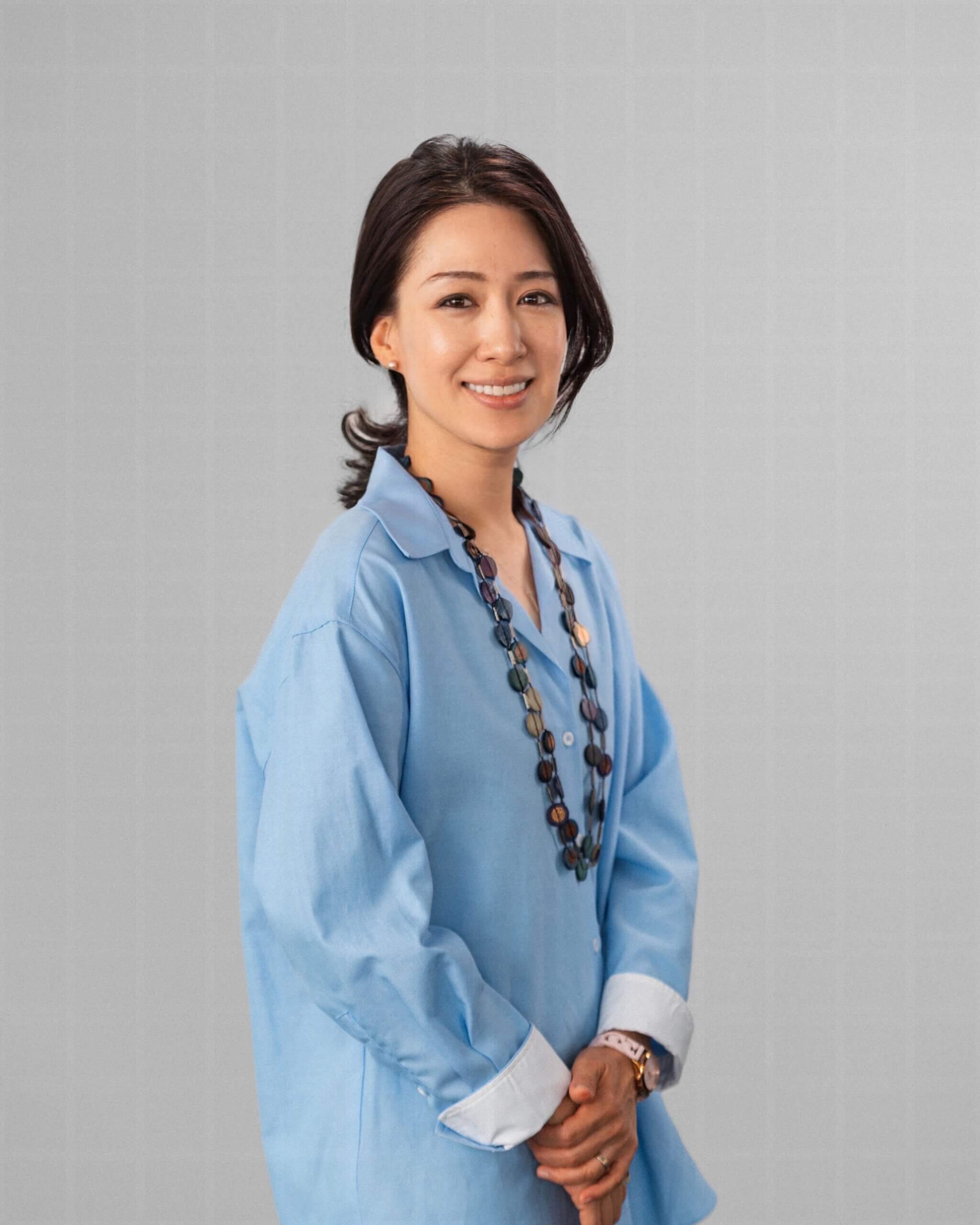 Jinnie Jeon, a woman with shoulder-length dark hair, wears a light blue button-up shirt and a beaded necklace, standing against a plain light grey background.