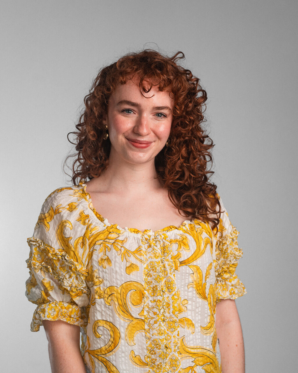 A woman with curly red hair wearing a white and yellow patterned dress stands in front of a plain gray background, smiling slightly at the camera.