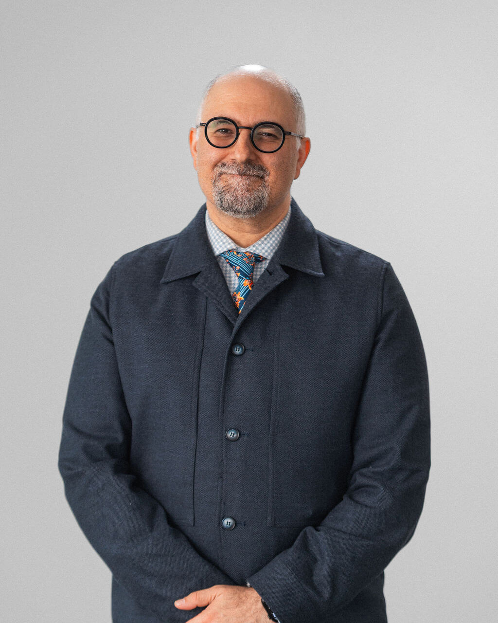 A middle-aged man, Amir Sepehry, with glasses, a beard, and a bald head stands in front of a plain light background, wearing a dark jacket, patterned shirt, and colorful tie.
