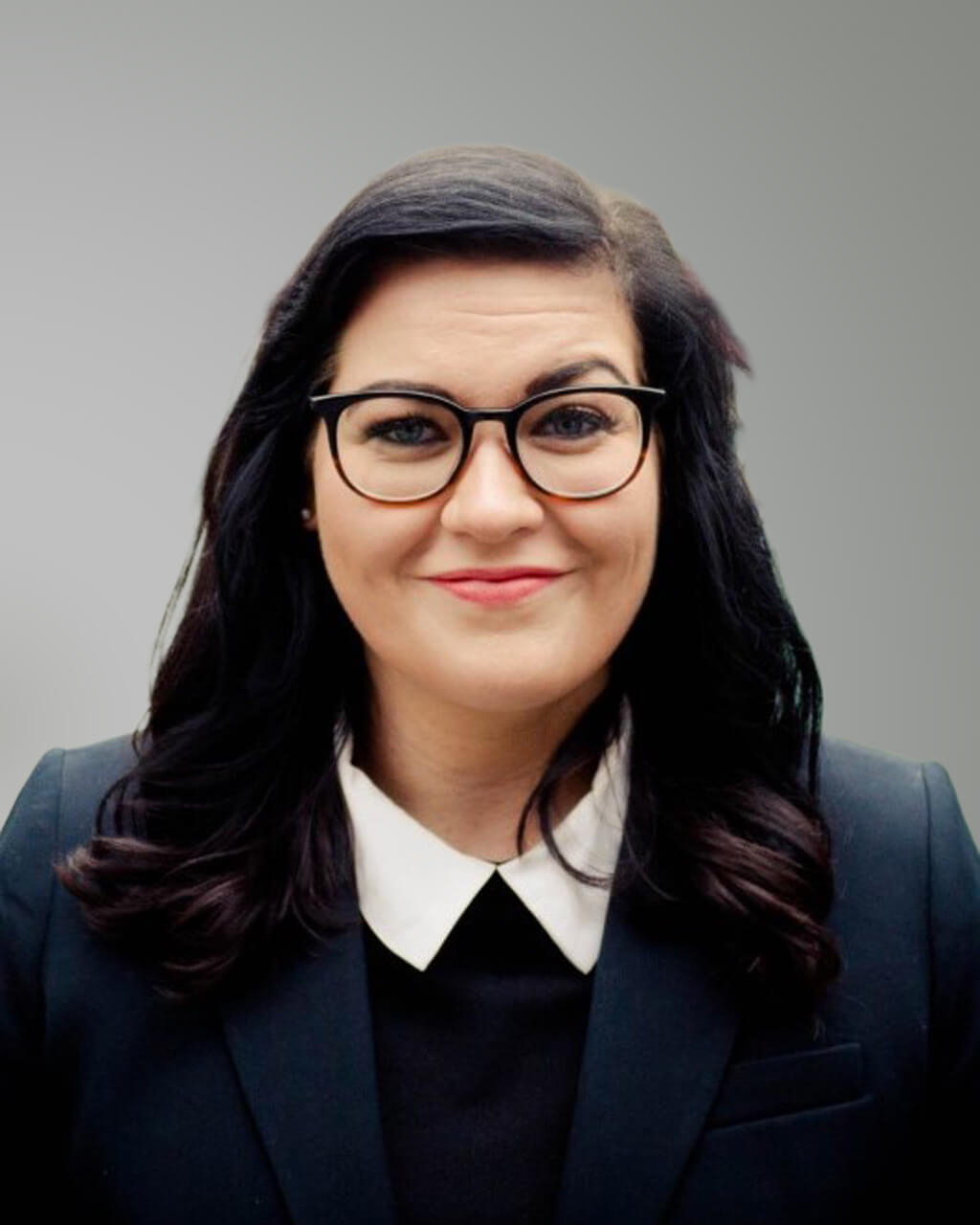 Ashlee Reed, a woman with long dark hair, wearing glasses, a black blazer, and a white-collared shirt, poses against a plain gray background.
