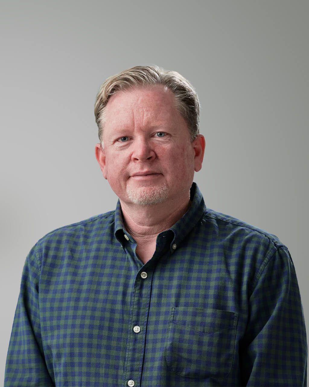 A middle-aged man with short, light hair and a trimmed beard wears a green and blue plaid shirt, standing against a plain gray background. He resembles Damon Elgie in his relaxed yet confident pose.