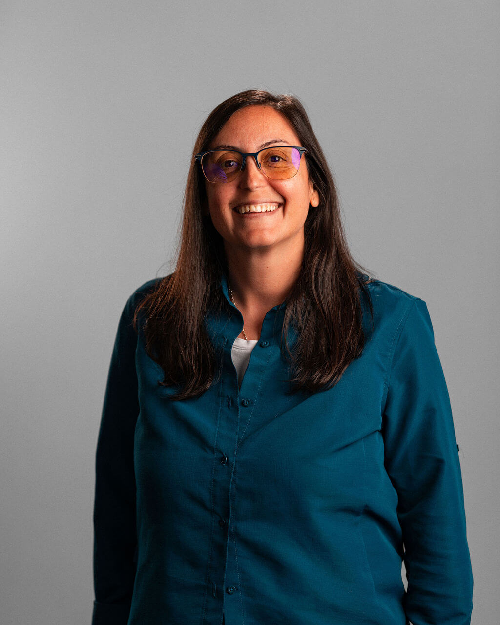 Elvita Kondili, with long dark hair and glasses, stands smiling in a teal button-up shirt against a plain gray background.