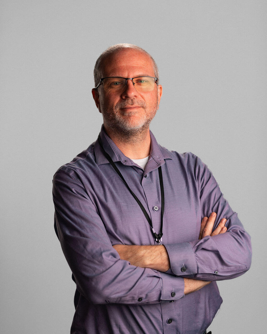 Jacob Kaiserman, a middle-aged man with glasses and a beard, stands with arms crossed, wearing a light purple button-up shirt and a black lanyard against a plain gray background.