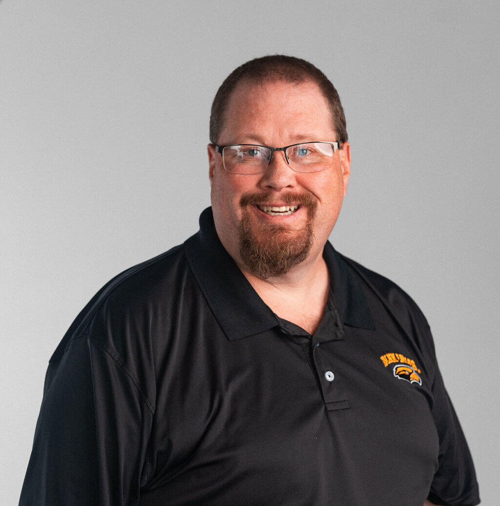 A man wearing glasses and a black polo shirt with a yellow and black Madson logo stands smiling against a plain gray background.