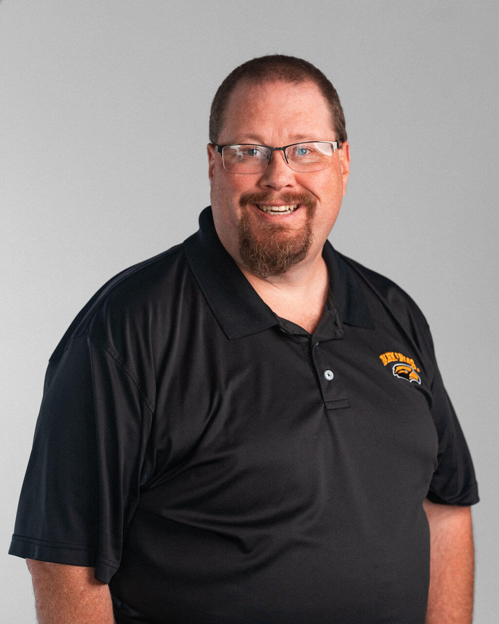 A man wearing glasses and a black polo shirt with a yellow and black Madson logo stands smiling against a plain gray background.