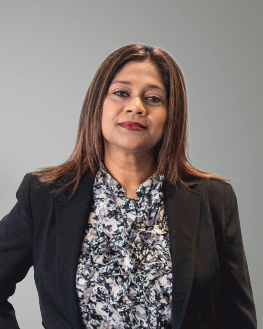 Hawwa Shiuna, a woman with straight brown hair, wears a black blazer and a floral blouse as she stands in front of a plain gray background.