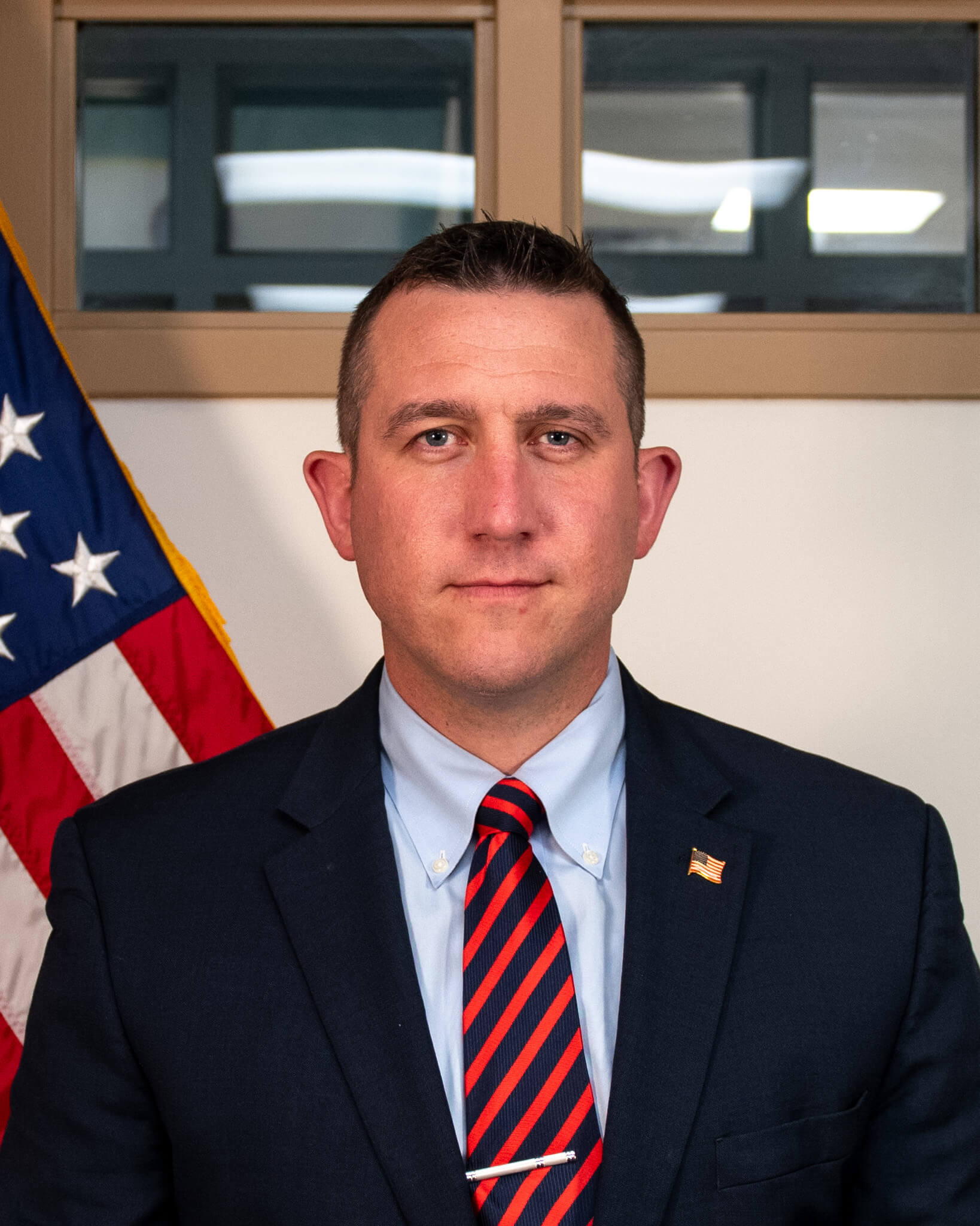 A man in a suit and striped tie stands in front of a U.S. flag and window, facing the camera with a neutral expression, reflecting leadership and commitment to issues like military mental health.