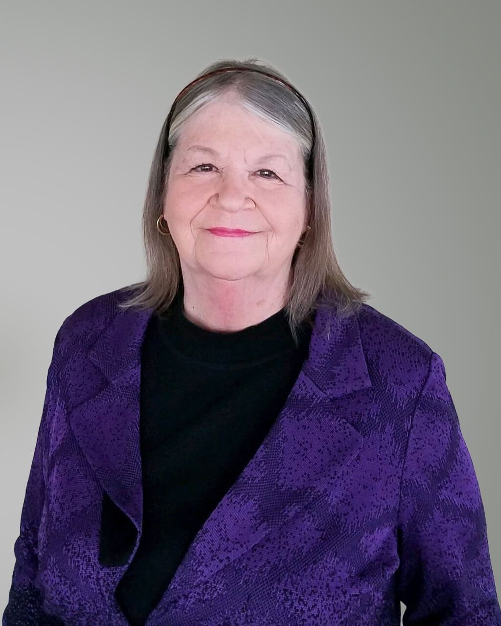 Sandra Kakacek, una mujer mayor de cabello canoso, lleva una chaqueta con estampado morado sobre un top negro mientras posa ante un fondo gris liso.