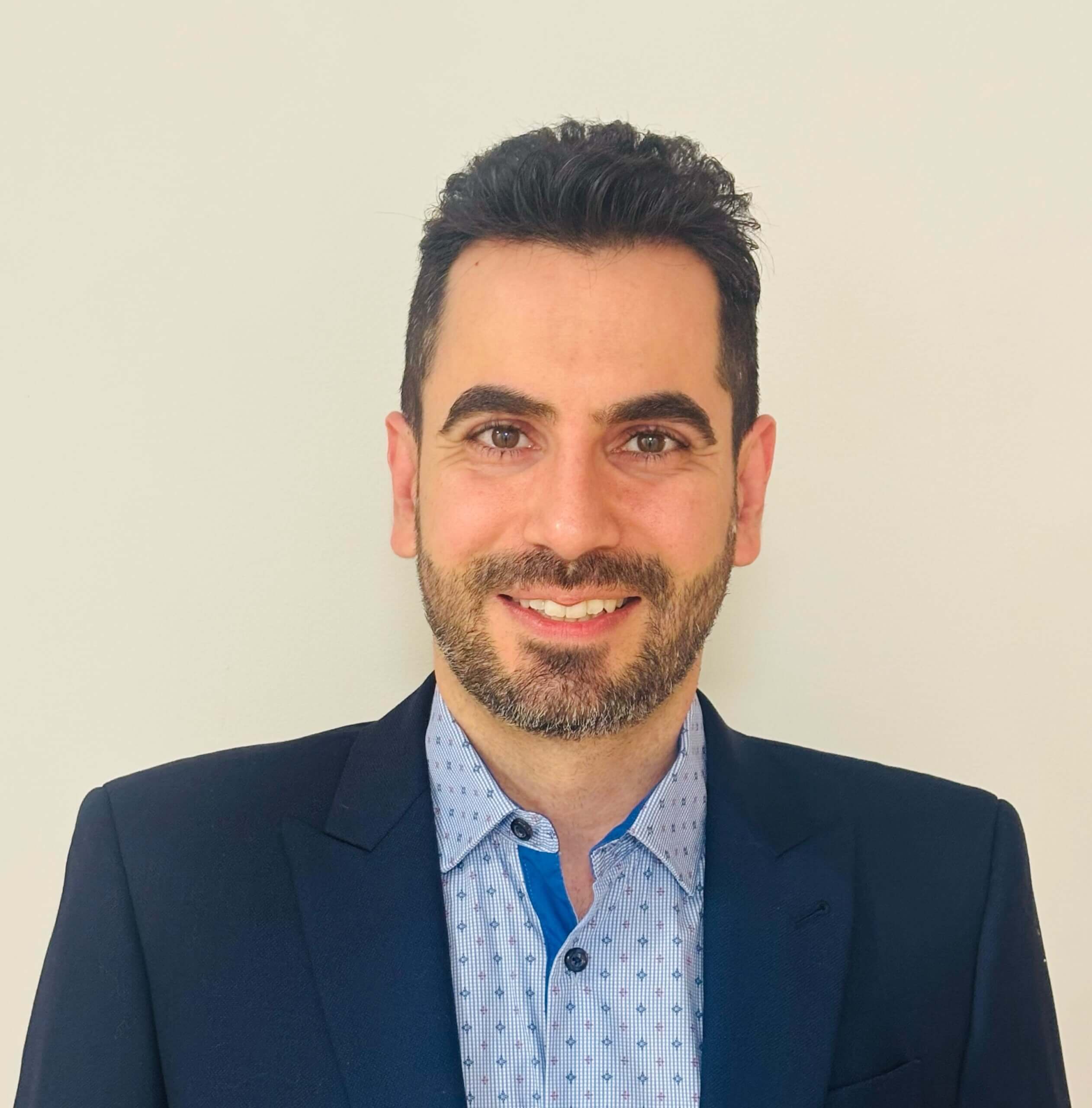 A man with dark hair and a beard is smiling at the camera, wearing a navy blazer and a light blue patterned shirt—this graduate speaker embodies a journey from refugee to psychologist against a plain light background.