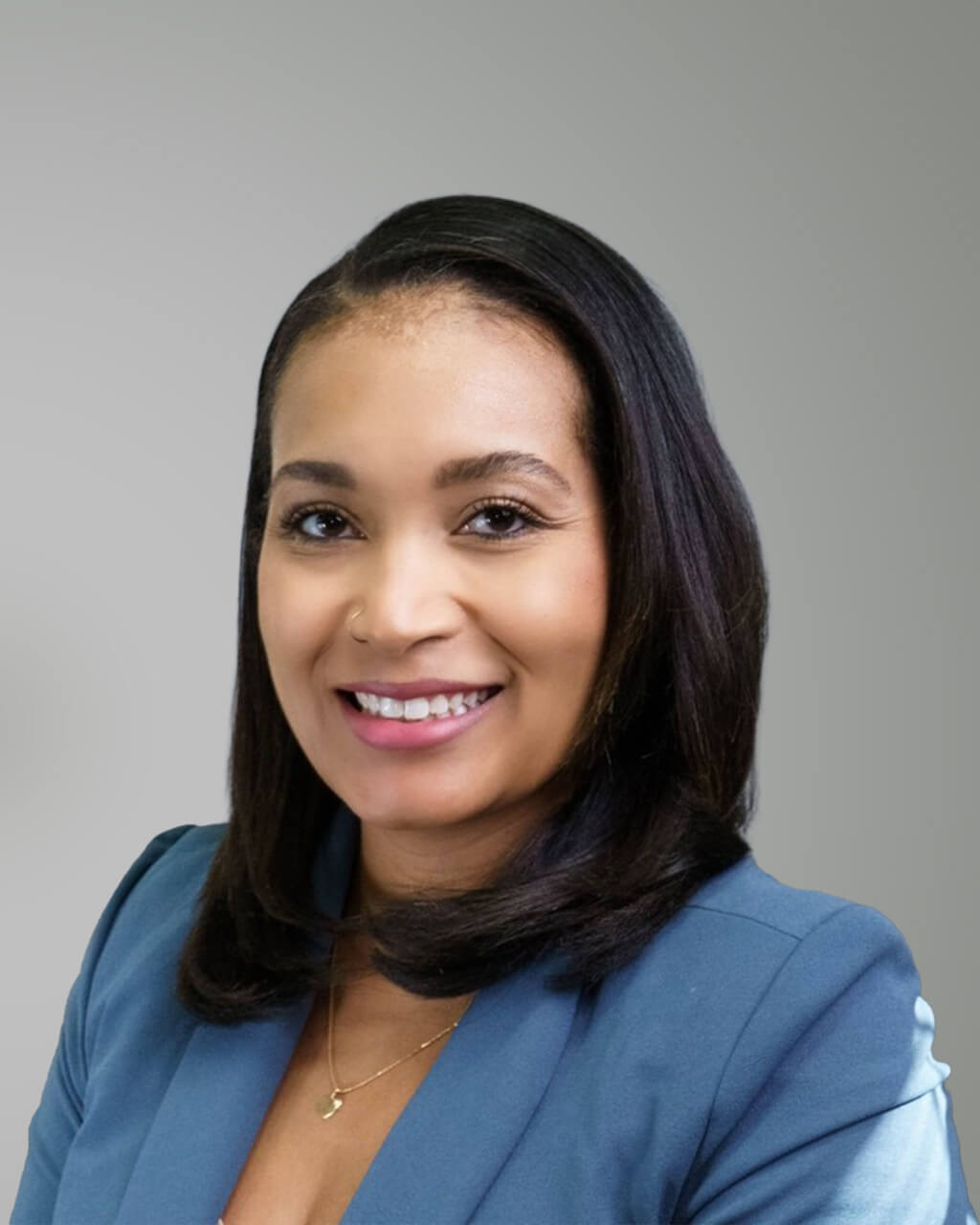 Shavonne, with straight black hair and a blue blazer, smiles in front of a plain light gray background.