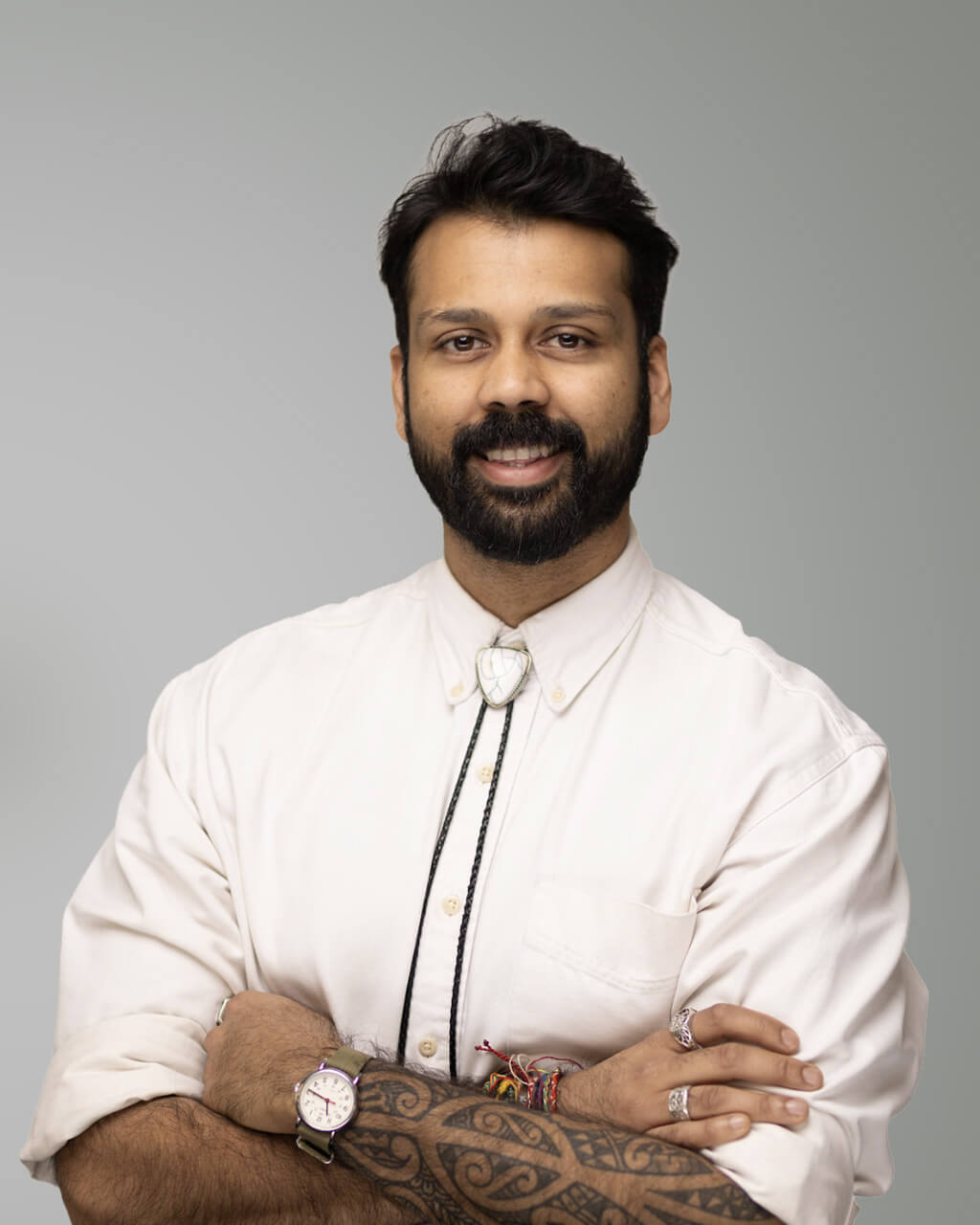 A man with a beard and mustache stands with arms crossed, wearing a light-colored shirt, bolo tie, wristwatch, rings, and bracelets, against a plain gray background.