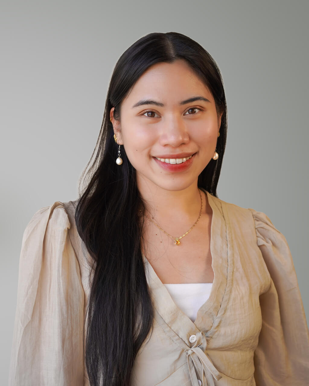 Bona Mikaela, a woman with long dark hair, wearing a beige blouse, white top, and pearl earrings, smiles at the camera against a plain light gray background.