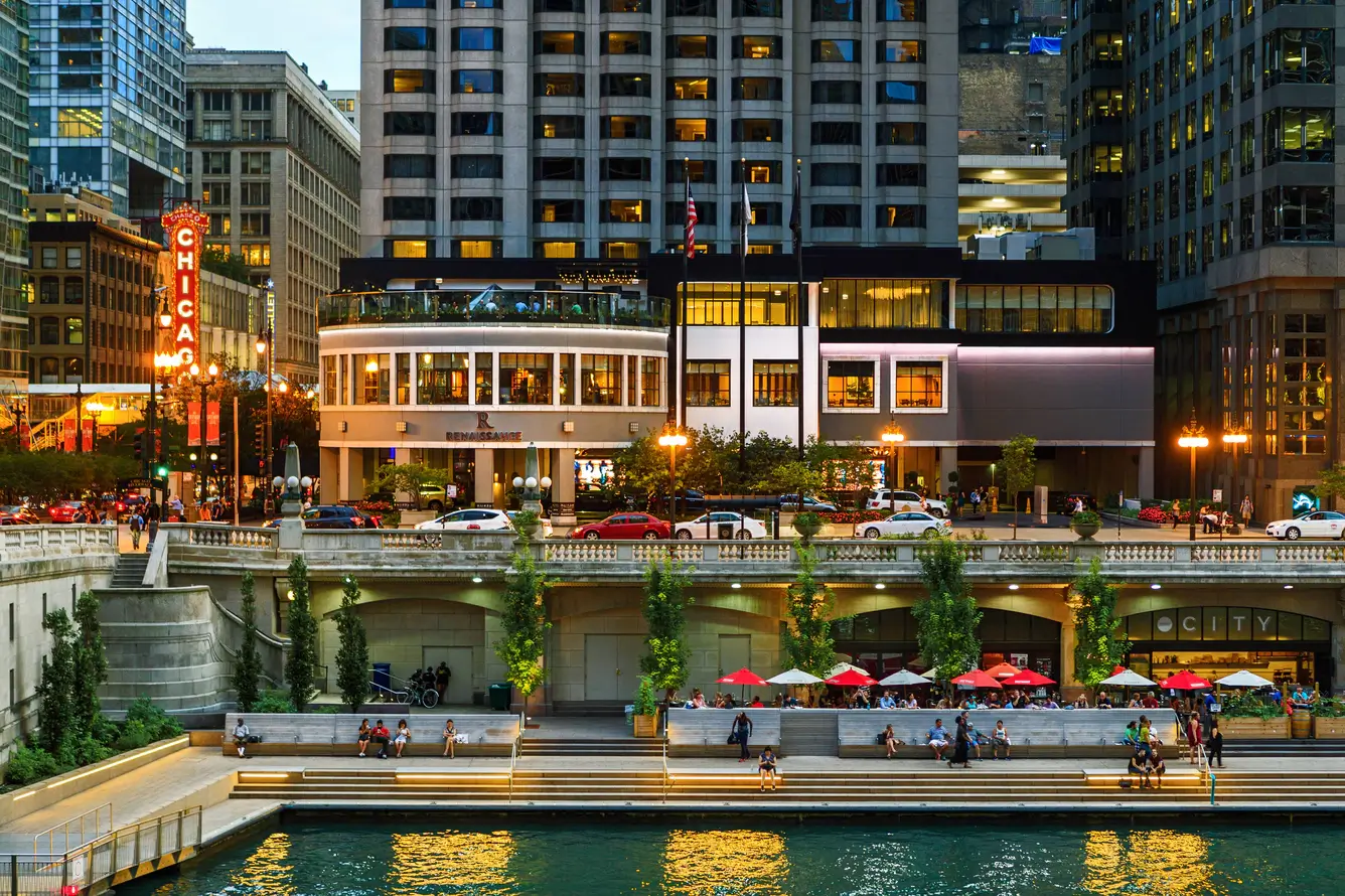 Les gens s'assoient et se promènent le long de la Chicago Riverwalk, près des terrasses de café, tandis que les immeubles de la ville et l'enseigne lumineuse du Chicago Theatre forment une toile de fond animée, reflétant l'effervescence qui règne dans la ville en cette période de remises de diplômes.