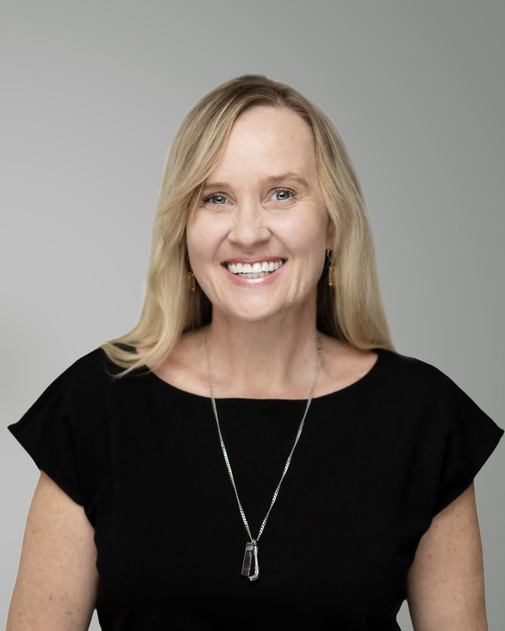 Erica Bearss, une femme aux longs cheveux blonds, porte un haut noir et un collier ; elle sourit à la caméra devant un fond uni gris clair.