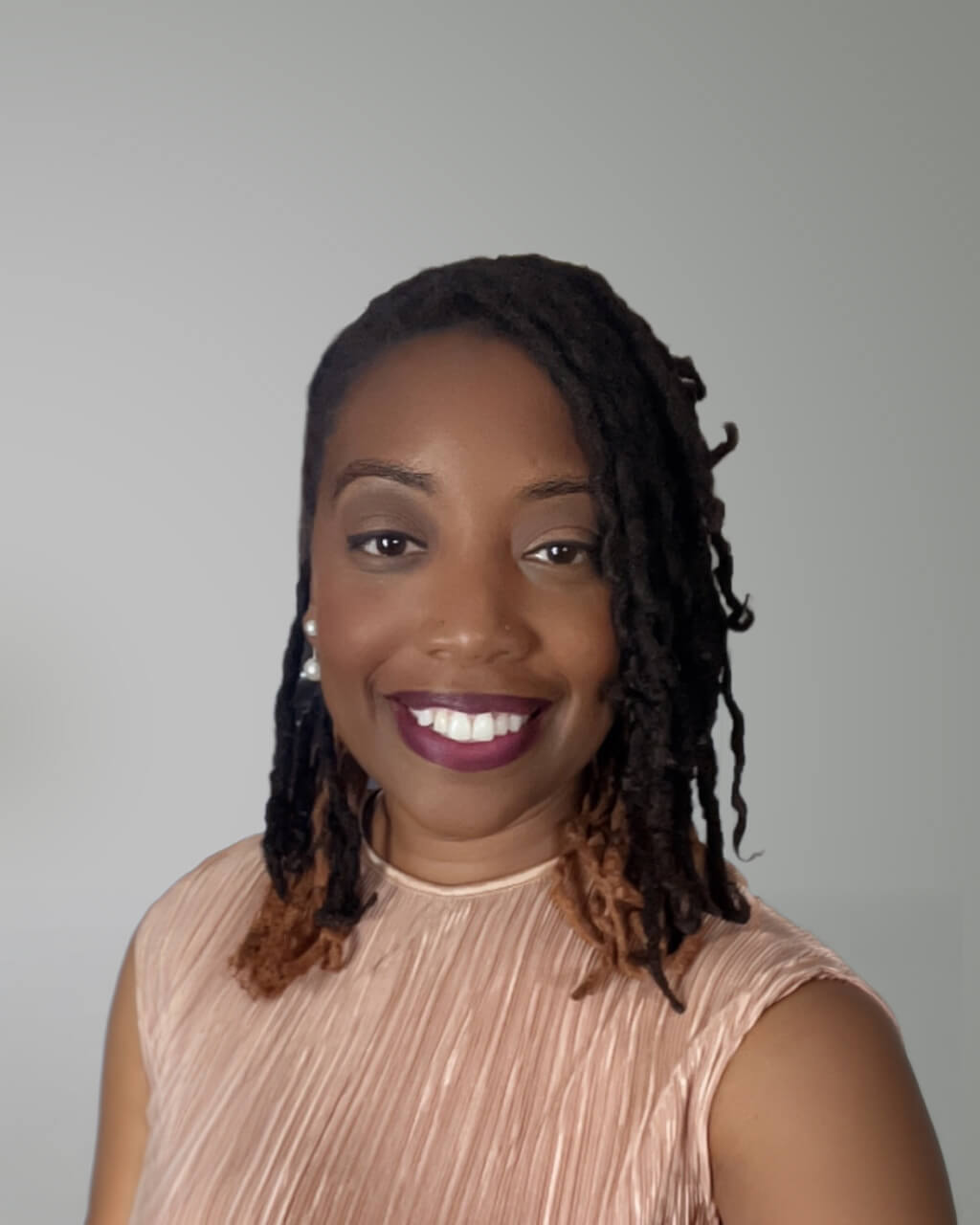 Jessica Fripp, a smiling woman with shoulder-length locs, wears a sleeveless, light-colored top against a plain gray background.