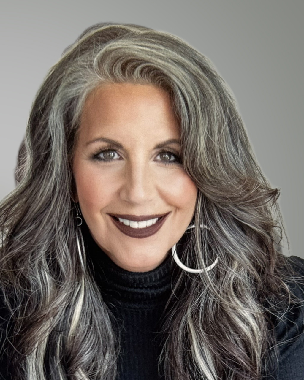 Smiling woman, Jennifer La Civita, with long wavy gray and brown hair, wearing a black turtleneck sweater and large hoop earrings, set against a plain gray background.