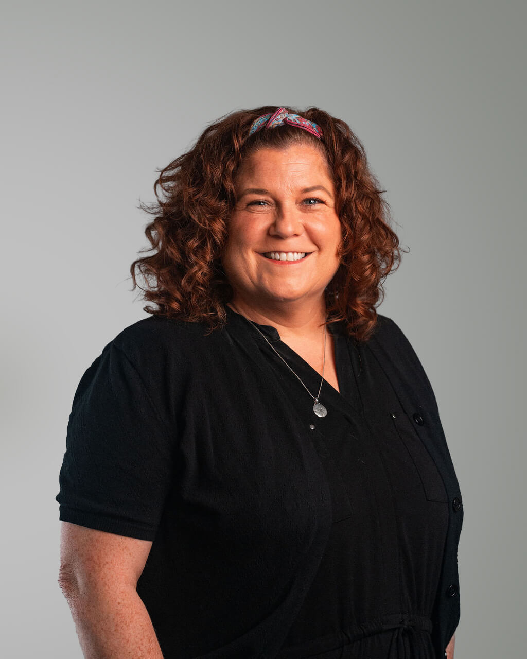 Mary, with curly brown hair, wearing a black outfit and a headband, smiles at the camera against a plain gray background—looking relaxed after finishing her laundry.