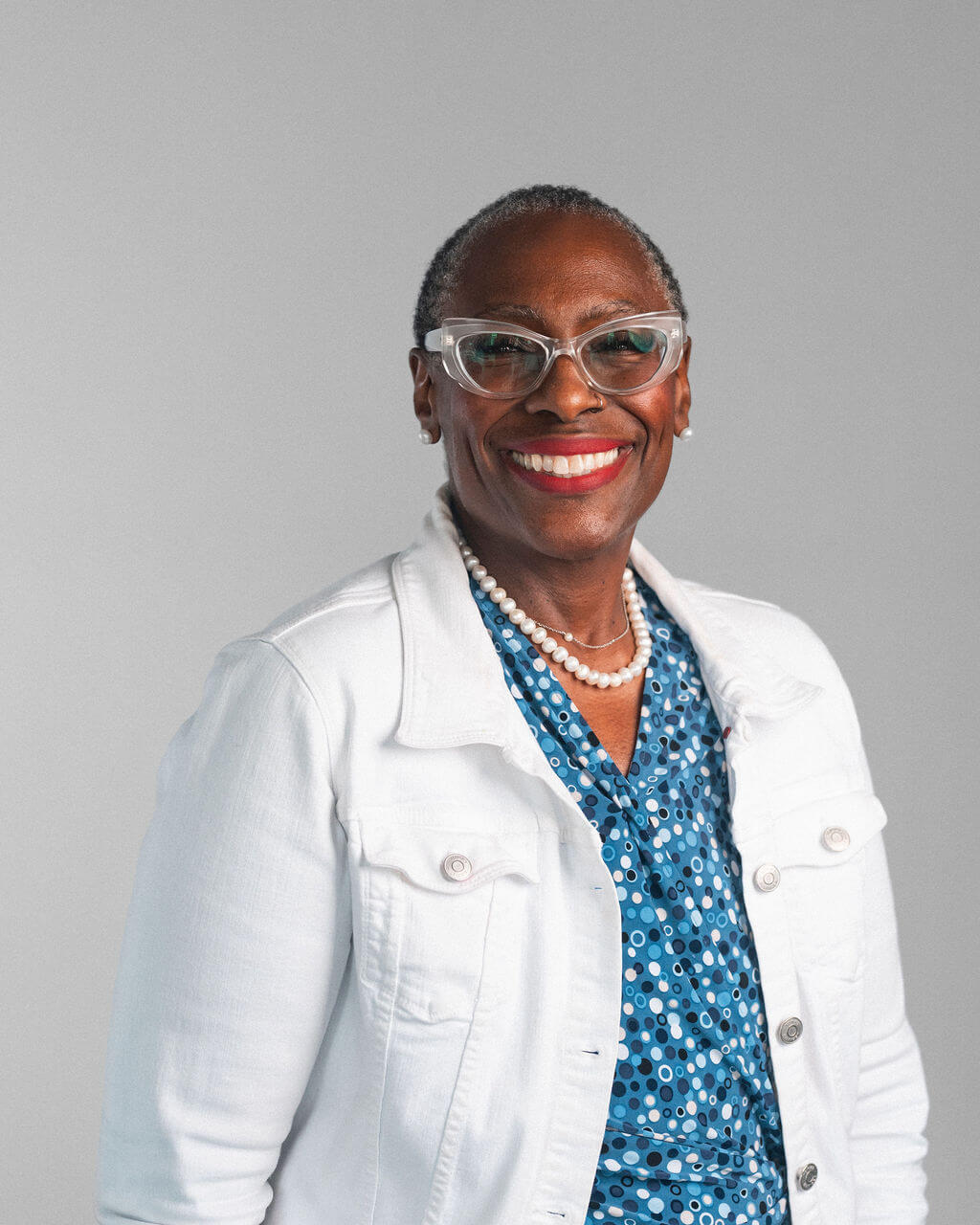 Monica Boyd-Layne, an older woman wearing glasses, a white jacket, a blue patterned blouse, and a pearl necklace, smiles warmly while standing against a plain gray background.