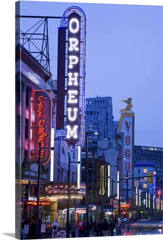 Neon signs for Orpheum, Café Crêpe, and Vogue light up a city street at dusk as people gather, some celebrating a graduation ceremony, with buildings glowing in the background.
