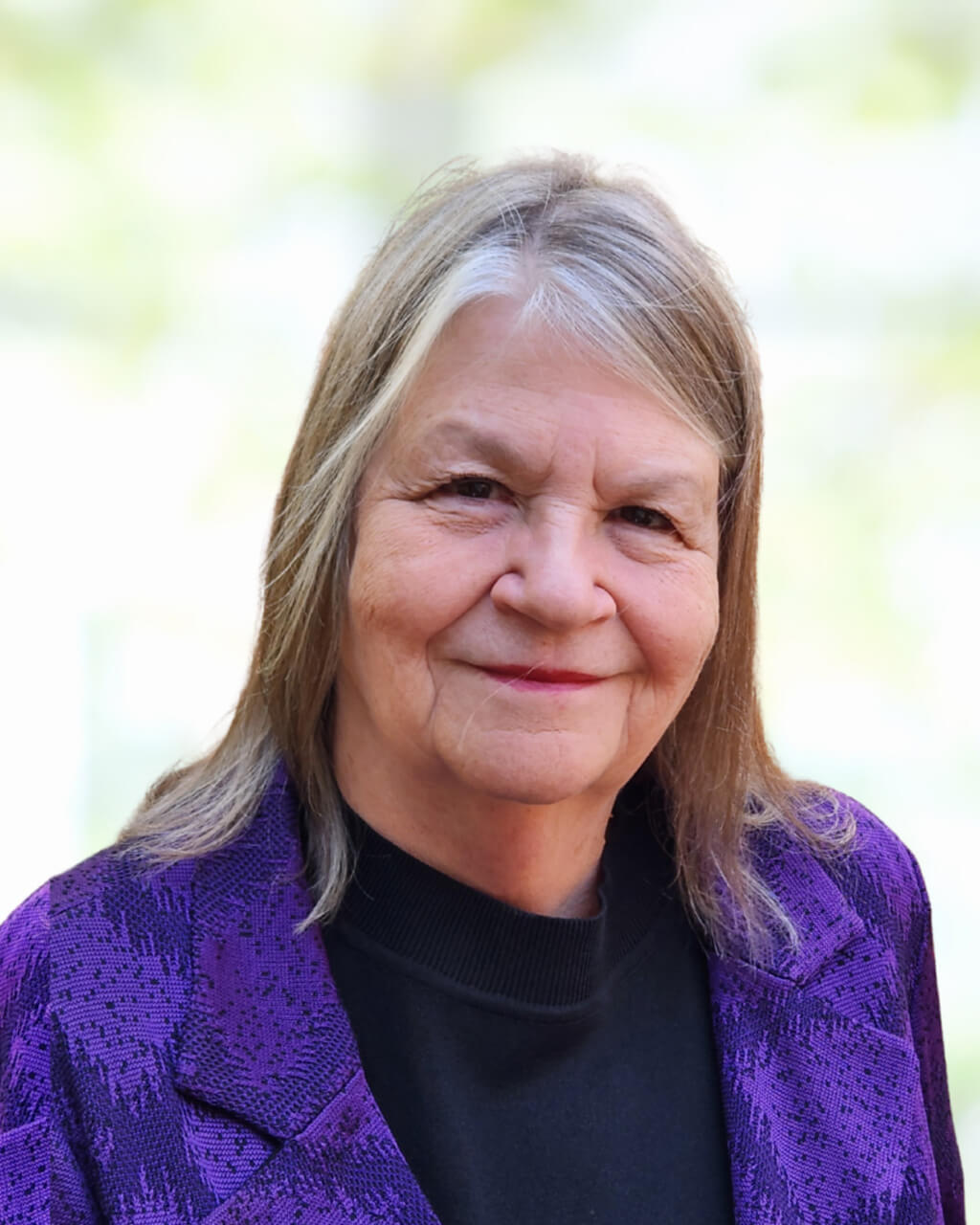 Sandra Kakacek, an older woman with straight gray hair, wearing a black top and a purple patterned jacket, smiles at the camera against a blurred light background.