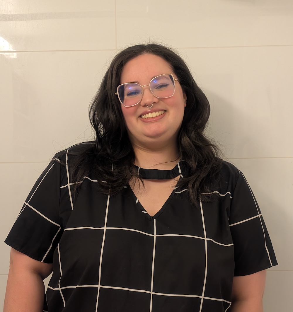 A person with long dark hair and glasses, wearing a black top with a white grid pattern, stands smiling with empathy against a plain tiled background.