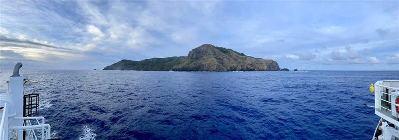 View of a rocky, green island in the distance surrounded by deep blue ocean, with parts of a ship visible on both sides—evoking the feeling of sailing to the edge of the world beneath a cloudy sky in search of meaning.