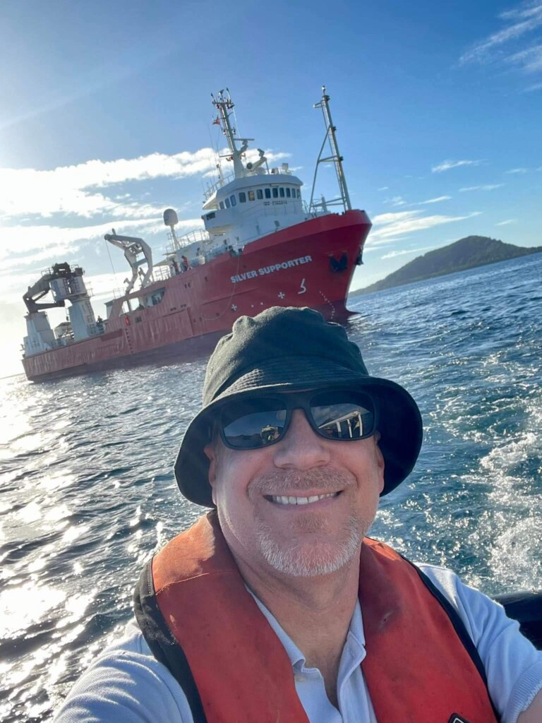 A man wearing a black hat and orange life vest smiles at the camera on a boat, with a large red research vessel and an island in the background, capturing the meaning of adventure at the edge of the world.