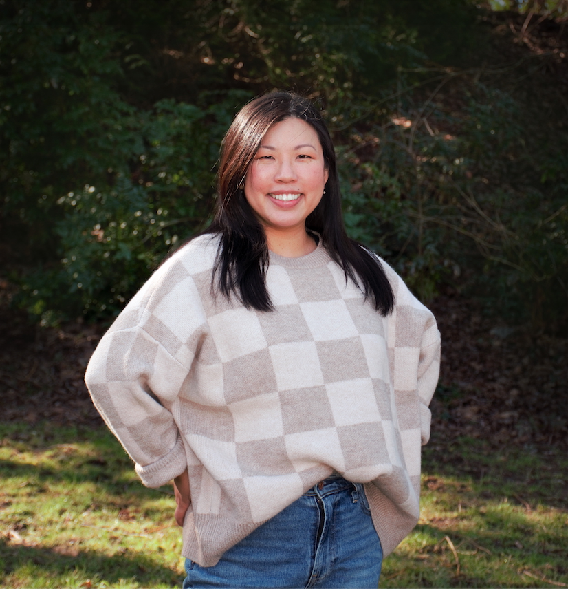 A woman stands outdoors on grass, smiling, wearing a beige and white checkered sweater and blue jeans. An Adler alum, she radiates positivity amid trees and bushes in the background.
