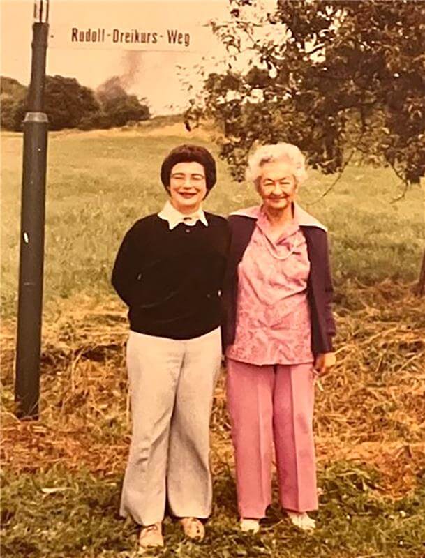 Two women stand side by side outdoors on a grassy path near a sign labeled "Rudolf-Dreikurs-Weg." Trees and an open field are visible in the background, highlighting the legacy of Ph.D. Eva Dreikurs Ferguson.