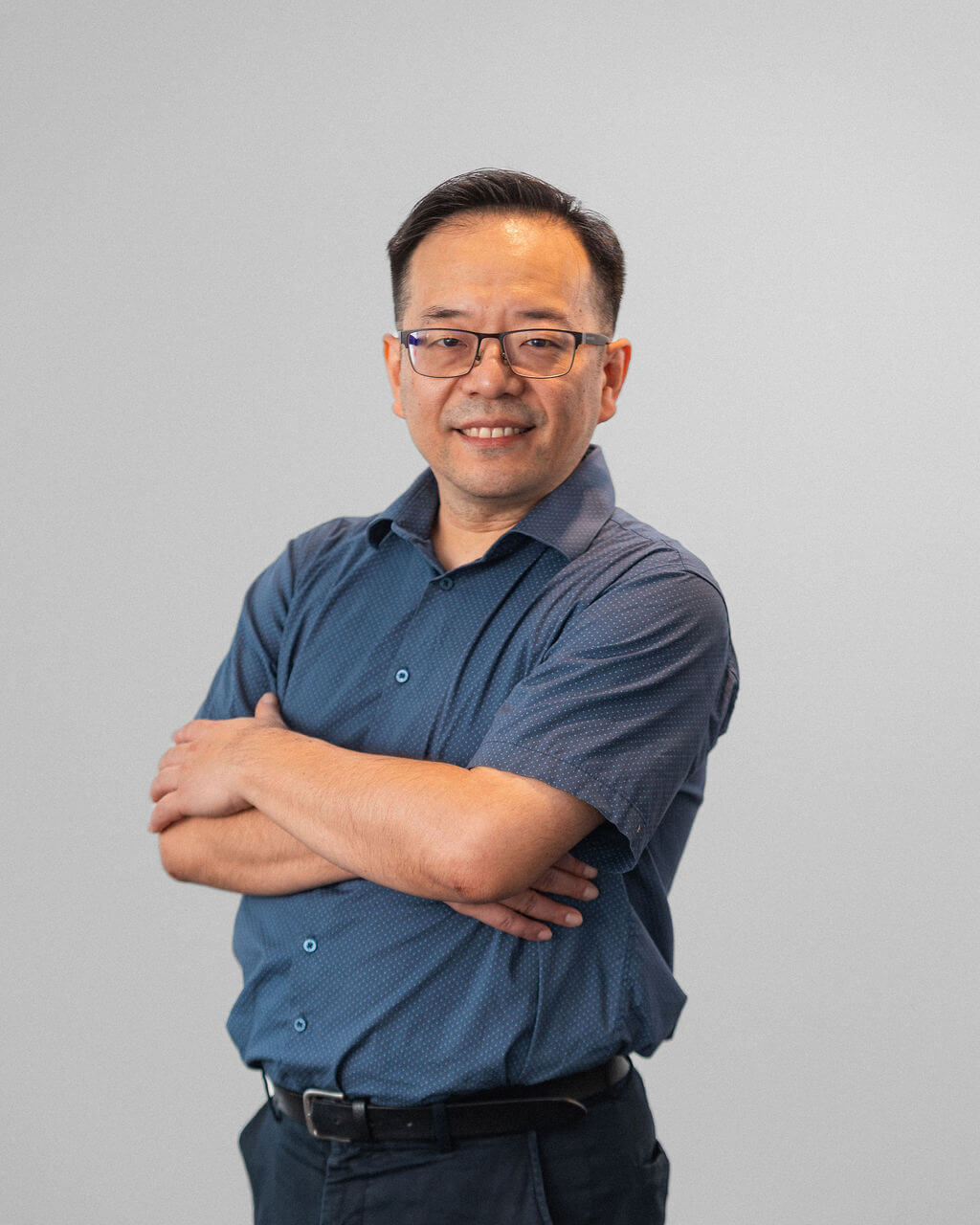 A man with short dark hair and glasses stands with arms crossed, wearing a blue short-sleeve button-up shirt and dark pants, against a plain light gray background.