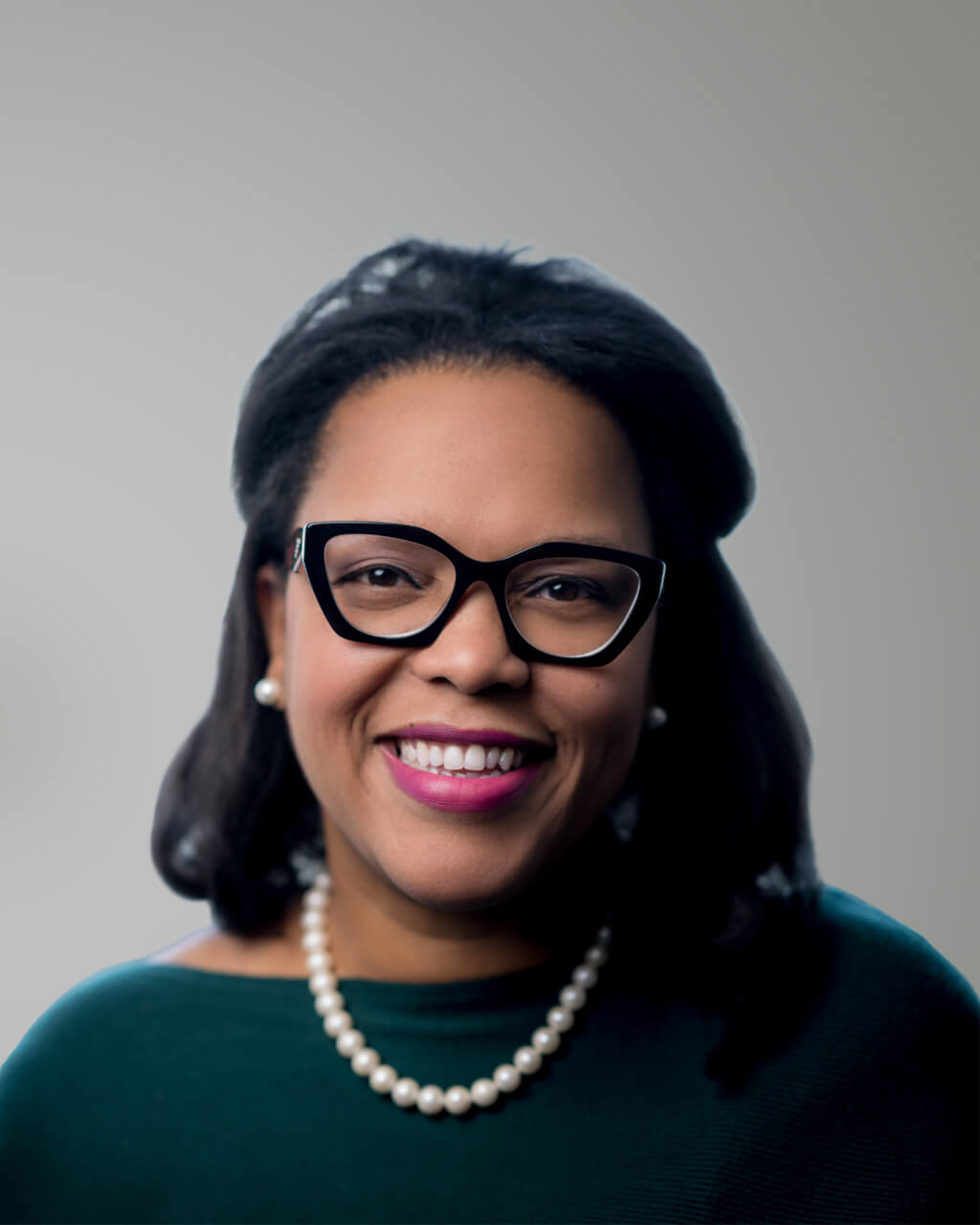 Una mujer de cabello oscuro, con gafas negras, un collar de perlas y un top oscuro, sonríe a la cámara ante un fondo liso: así se presenta Kesha S. Burch.