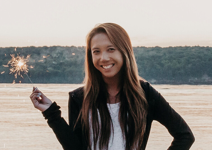 A doctoral student stands by the water holding a lit sparkler, smiling at the camera, with trees and a light sky in the background—a joyful moment inspired by the Tiffany Gee Memorial Scholarship.
