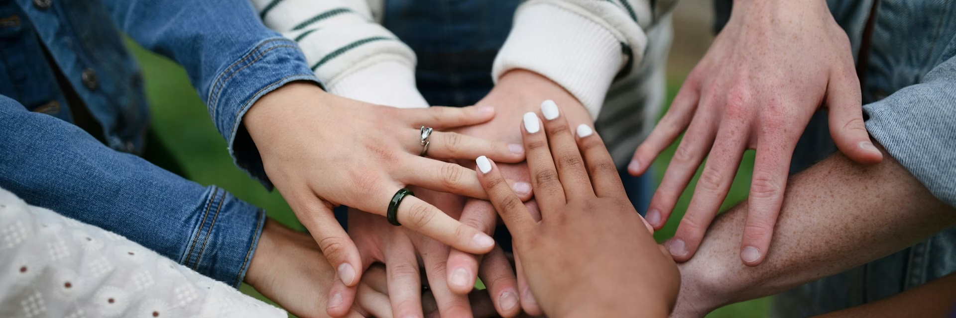 A group of people stand in a circle and stack their hands together in the center, showing teamwork and unity—the price of belonging embraced by all.