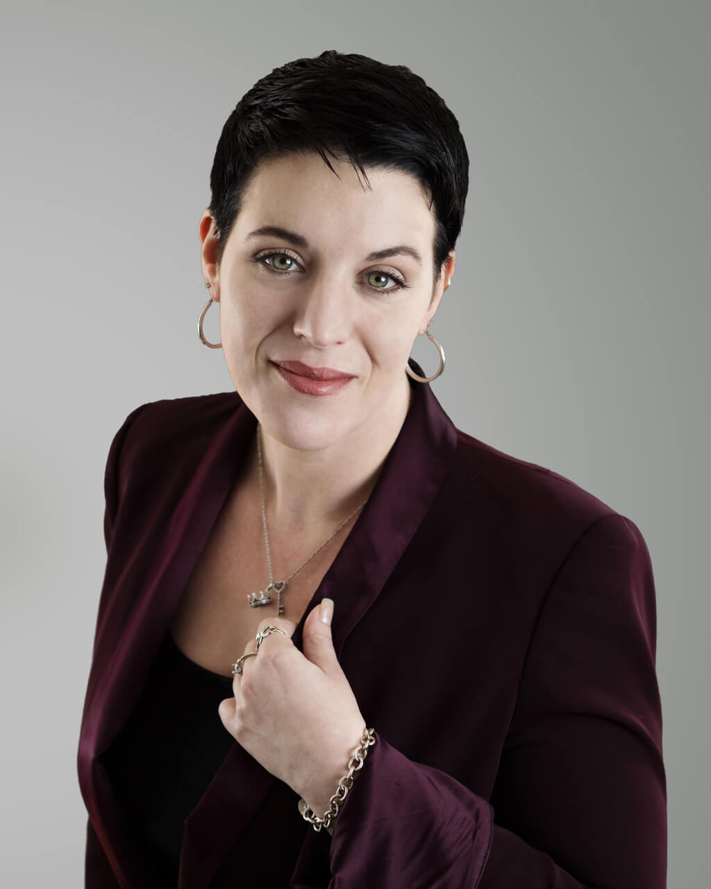 Karen Caffee, a woman with short dark hair, stands confidently in front of a plain gray background. She wears a burgundy blazer over a black top, complemented by silver jewelry and hoop earrings.