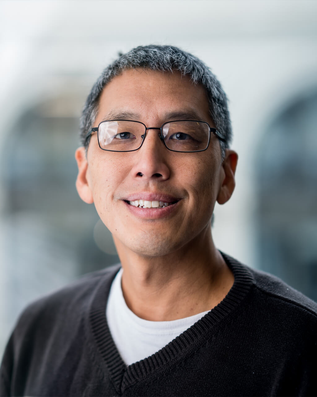 A middle-aged man with short gray hair and glasses is smiling, wearing a dark sweater over a white shirt, with a blurred indoor background.