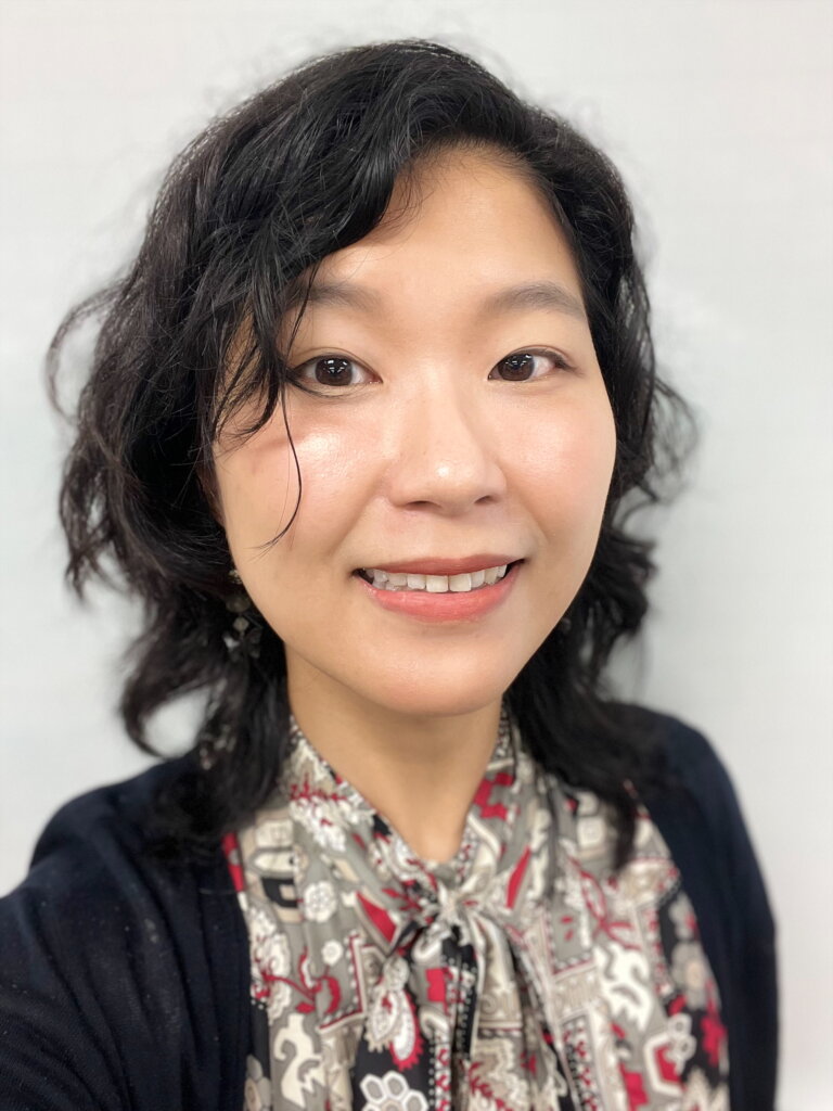 A doctoral student with wavy dark hair, wearing a patterned blouse and black cardigan, smiles at the camera against a plain light background. She is a proud recipient of the Tiffany Gee Memorial Scholarship.