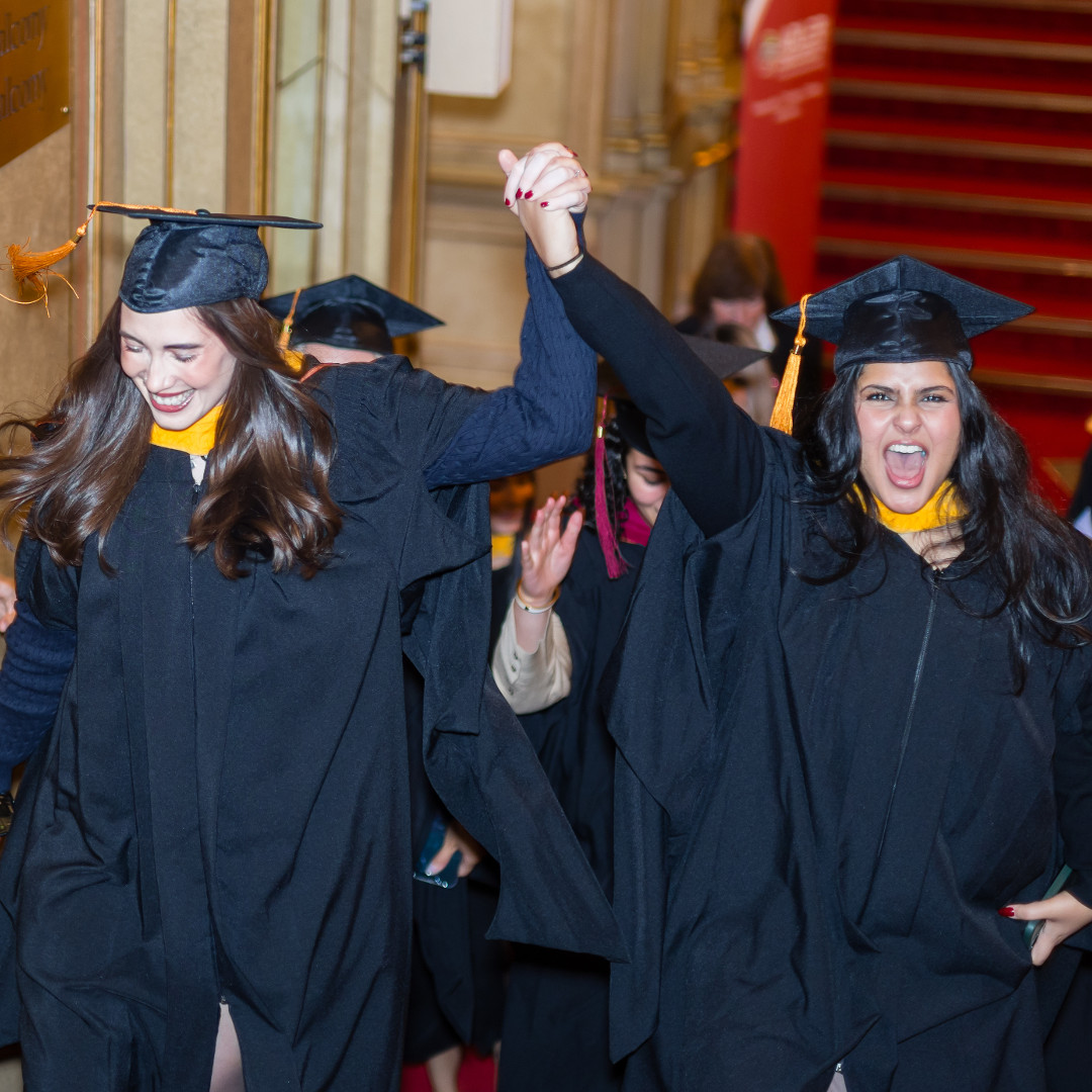 Two graduates in caps and gowns hold hands and cheer while walking up stairs at a graduation ceremony, celebrating their success at Adler University—recognized by Newsweek as one of the best colleges for women.