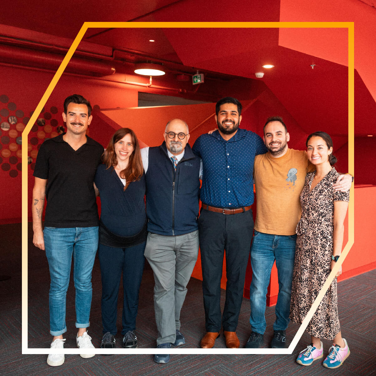 Six people stand side by side, smiling, in a brightly lit room with red walls and geometric decor during Founders’ Days.